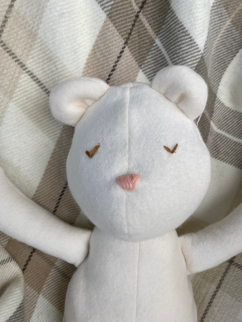 May include: White plush toy bear with embroidered eyes and a pink nose. The bear is lying on a plaid blanket.