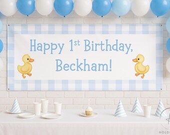 Puede incluir: Una pancarta de fiesta de cumpleaños con el texto "Happy 1st Birthday, Beckham!" en azul, enmarcada por un patrón de cuadros azules y blancos. La pancarta está decorada con ilustraciones de patos amarillos y rodeada de globos azules y blancos. Una mesa está puesta con artículos de fiesta.