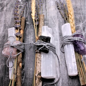 Könnte beinhalten: Drei Bündel aus Selenitkristallen, getrocknetem Lavendel und Weizenähren, gebunden mit grauem Garn. Jedes Bündel enthält einen anderen Kristall: Rosenquarz, schwarzer Turmalin und Amethyst. Der Hintergrund ist verwittertes graues Holz.
