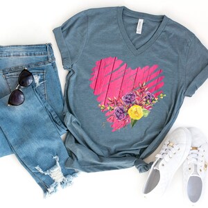 May include: A teal blue V-neck t-shirt with a pink heart design featuring a floral bouquet. The heart is made to look like weathered wood with pink paint. The bouquet includes purple, yellow, and green flowers.