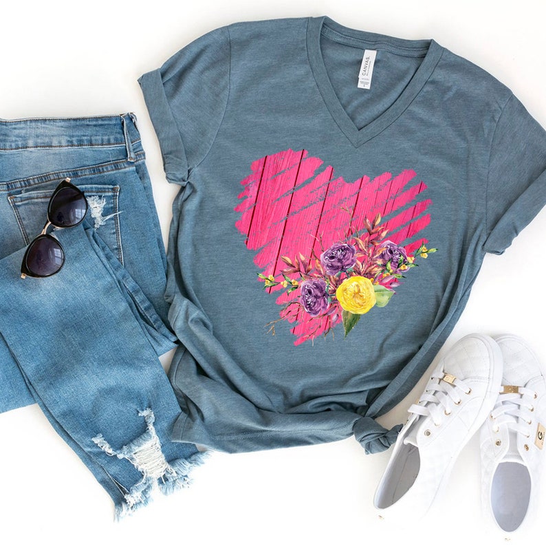 May include: A teal blue V-neck t-shirt with a pink heart design featuring a floral bouquet. The heart is made to look like weathered wood with pink paint. The bouquet includes purple, yellow, and green flowers.
