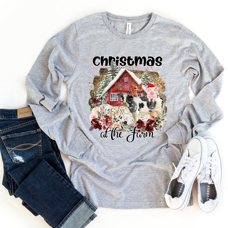 Puede incluir: Una camiseta gris de manga larga con el texto "Christmas at the Farm" en negro. El dise&ntilde;o presenta un granero rojo con una vaca, un cerdo y un gallo delante. El dise&ntilde;o est&aacute; rodeado de flores rojas y blancas y purpurina dorada.