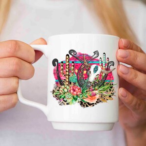May include: White ceramic mug with a colourful design featuring a skull, horseshoes, and cacti. The design has a pink, turquoise, and gold colour scheme.