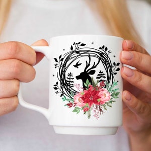 May include: White ceramic mug featuring a black silhouette of a deer head within a wreath of leaves and berries. The design includes a bird, pine trees, and a cluster of red and pink flowers. The mug has a white handle.