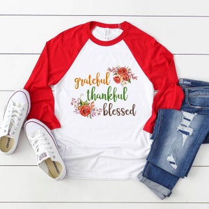 Puede incluir: Una camiseta de b&eacute;isbol blanca y roja con las palabras "grateful thankful blessed" impresas en la parte delantera. La camiseta est&aacute; decorada con un dise&ntilde;o floral en tonos naranja, rojo y verde.