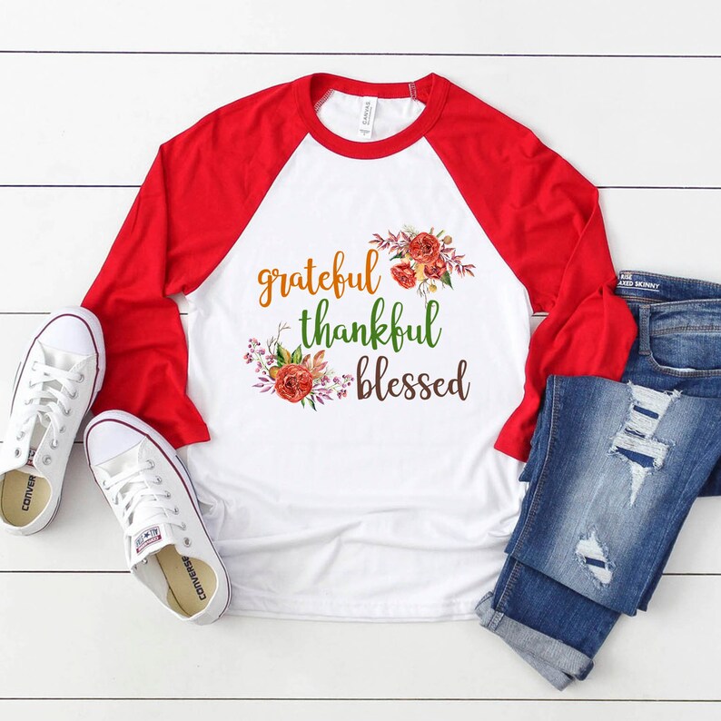 Puede incluir: Una camiseta de b&eacute;isbol blanca y roja con las palabras "grateful thankful blessed" impresas en la parte delantera. La camiseta est&aacute; decorada con un dise&ntilde;o floral en tonos naranja, rojo y verde.
