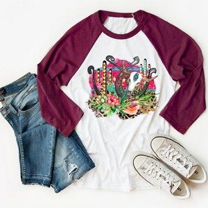 May include: A white and maroon baseball tee shirt featuring a graphic design of a skull, horseshoes, and cacti on a pink and blue background. The design is surrounded by leopard print and floral accents.