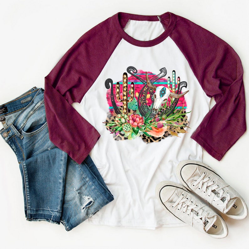 May include: A white and maroon baseball tee shirt featuring a graphic design of a skull, horseshoes, and cacti on a pink and blue background. The design is surrounded by leopard print and floral accents.