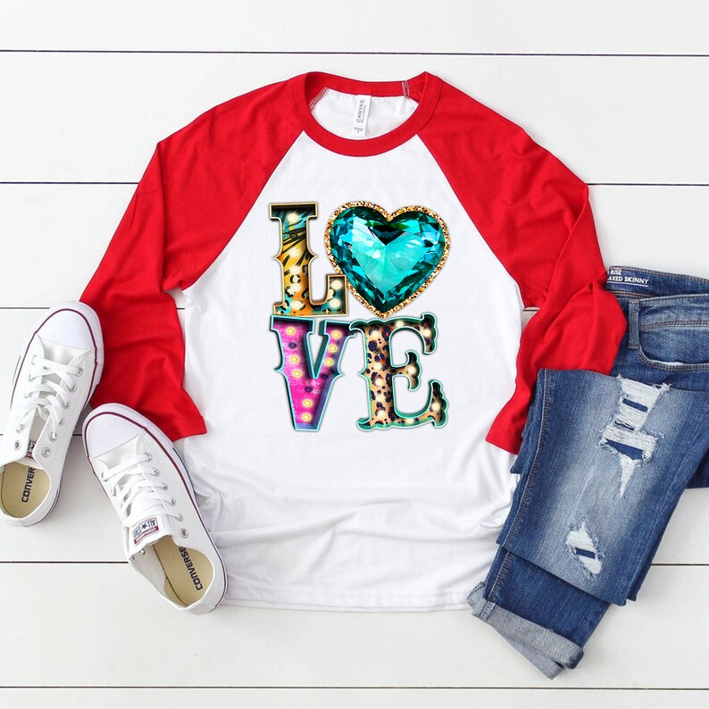Puede incluir: Una camiseta de b&eacute;isbol blanca y roja con un gr&aacute;fico de la palabra "LOVE" en letras de colores con un gran coraz&oacute;n turquesa en el medio. Las letras est&aacute;n decoradas con purpurina dorada y estampado de leopardo.