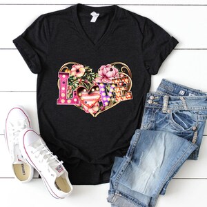 Puede incluir: Una camiseta negra de cuello en V con un gr&aacute;fico de la palabra "LOVE" en forma de coraz&oacute;n rodeada de flores. Las letras est&aacute;n decoradas con luces y un coraz&oacute;n rojo con un patr&oacute;n blanco y azul est&aacute; en el centro.