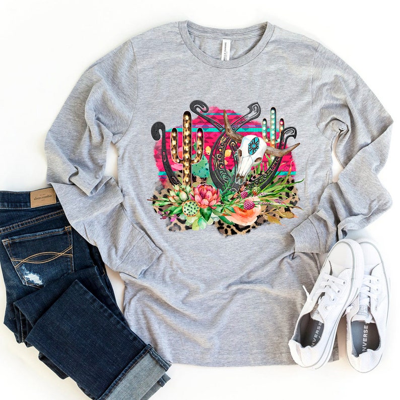 May include: Grey long-sleeved t-shirt with a graphic design of a skull, horseshoes, and cacti in a pink, turquoise and gold colour scheme.