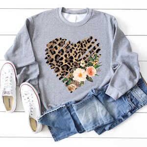 May include: A gray sweatshirt with a heart-shaped design featuring a leopard print and floral accents. The heart is outlined with a distressed, ripped effect.