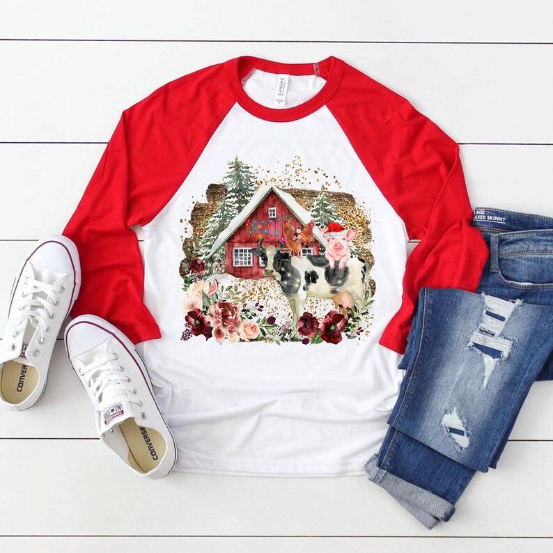 Pu&ograve; includere: Una maglietta da baseball bianca e rossa con un design a tema natalizio che presenta un fienile rosso, una mucca, un maiale e un arrangiamento floreale. Il design &egrave; su uno sfondo bianco con glitter dorato.