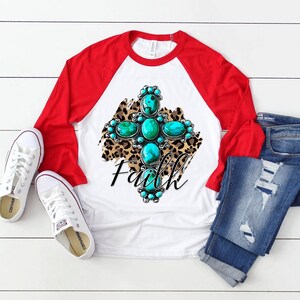 Puede incluir: Una camiseta de b&eacute;isbol blanca y roja con un dise&ntilde;o de cruz turquesa y plata sobre un fondo de estampado de leopardo. La palabra "Faith" est&aacute; escrita debajo de la cruz.