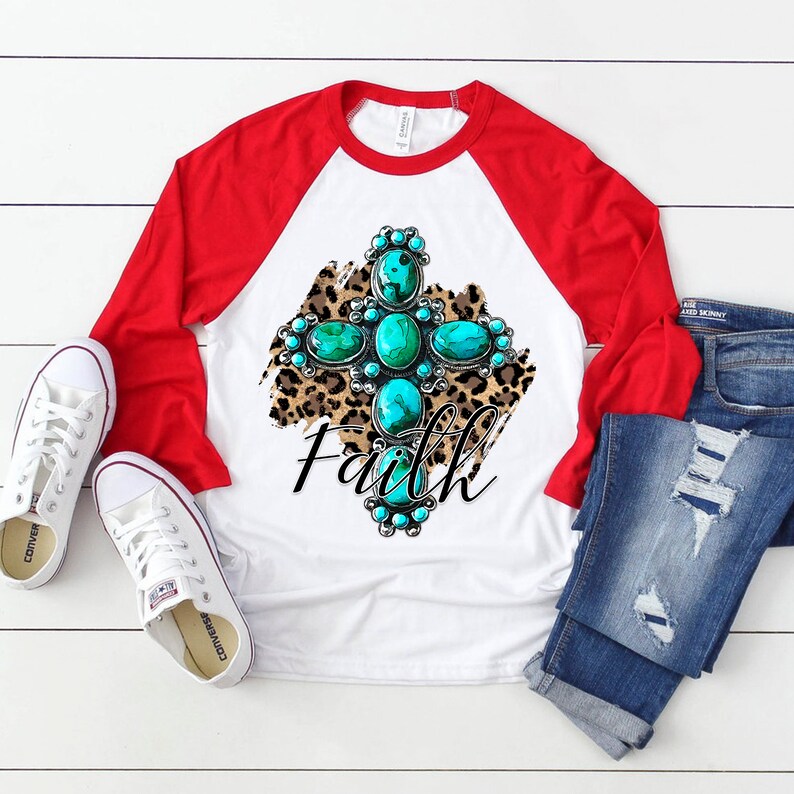 Puede incluir: Una camiseta de b&eacute;isbol blanca y roja con un dise&ntilde;o de cruz turquesa y plata sobre un fondo de estampado de leopardo. La palabra "Faith" est&aacute; escrita debajo de la cruz.