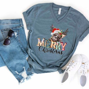 Pu&ograve; includere: Una maglietta blu petrolio con scollo a V con una grafica di un bulldog francese che indossa un cappello di Babbo Natale e corna di renna. Il testo "Merry Christmas" &egrave; scritto in lettere colorate con lampadine intorno.