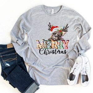Pu&ograve; includere: Una maglietta grigia a maniche lunghe con un design festivo che presenta un bulldog francese con un cappello di Babbo Natale e corna di renna. Il testo "Merry Christmas" &egrave; visualizzato in lettere colorate con lampadine.