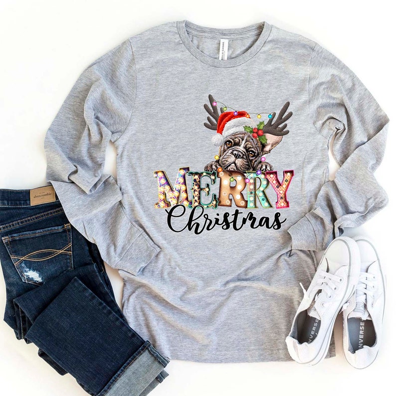 Pu&ograve; includere: Una maglietta grigia a maniche lunghe con un design festivo che presenta un bulldog francese con un cappello di Babbo Natale e corna di renna. Il testo "Merry Christmas" &egrave; visualizzato in lettere colorate con lampadine.