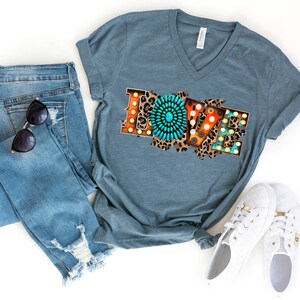 May include: A teal blue V-neck t-shirt with a graphic design of the word "LOVE" in colorful, distressed letters with a turquoise and brown circular design in the center of the "O".