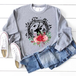 May include: A gray sweatshirt featuring a deer head silhouette within a black wreath, accented with red and pink floral elements. The design is paired with blue jeans and white canvas sneakers, creating a casual, everyday look.