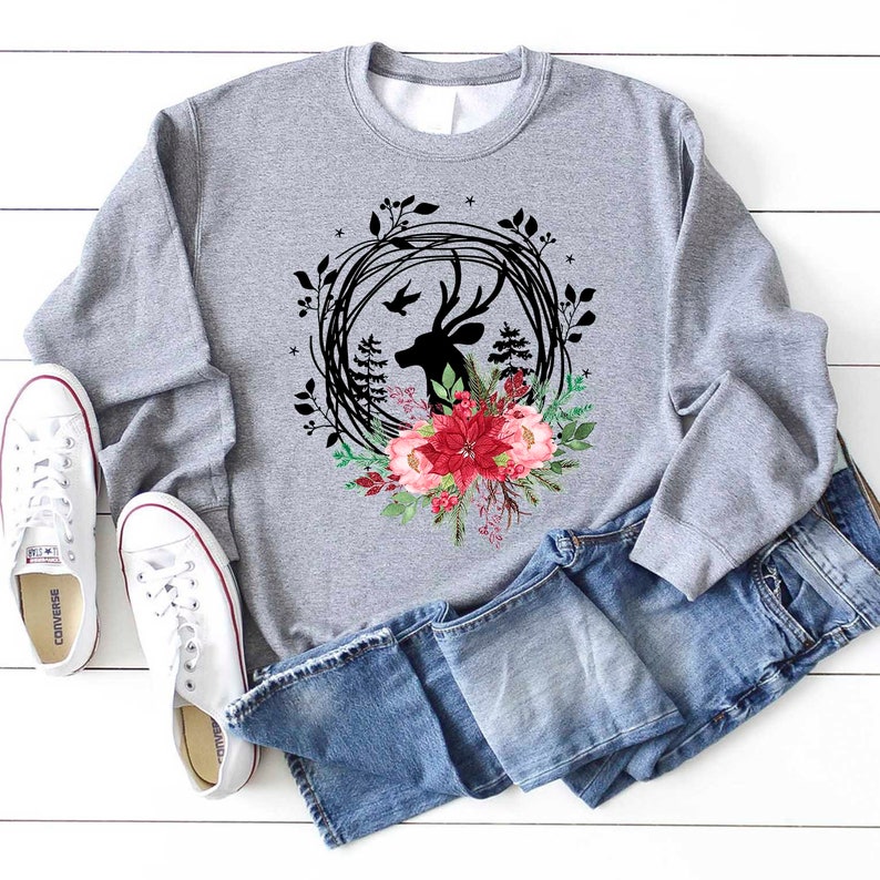May include: A gray sweatshirt featuring a deer head silhouette within a black wreath, accented with red and pink floral elements. The design is paired with blue jeans and white canvas sneakers, creating a casual, everyday look.
