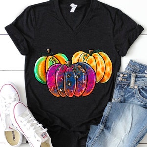 Peut inclure: Un t-shirt noir avec un motif color&eacute; de trois citrouilles. Les citrouilles sont peintes dans des tons d'orange, de vert, de rose et de bleu. Le motif a un effet de peinture &eacute;clabouss&eacute;e.