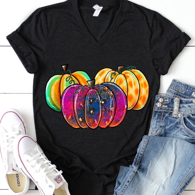 Peut inclure: Un t-shirt noir avec un motif color&eacute; de trois citrouilles. Les citrouilles sont peintes dans des tons d'orange, de vert, de rose et de bleu. Le motif a un effet de peinture &eacute;clabouss&eacute;e.
