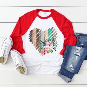 May include: A white and red baseball tee shirt with a distressed wood heart design and floral accents. The heart is made of wood planks in shades of brown, blue, and green. The floral accents are pink, purple, and white.