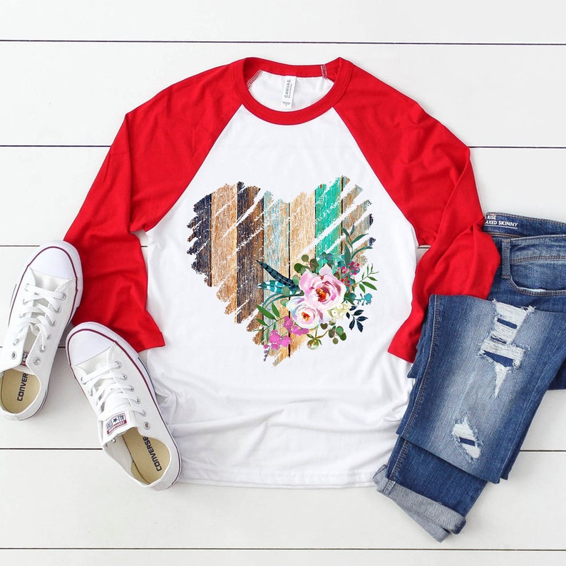 May include: A white and red baseball tee shirt with a distressed wood heart design and floral accents. The heart is made of wood planks in shades of brown, blue, and green. The floral accents are pink, purple, and white.