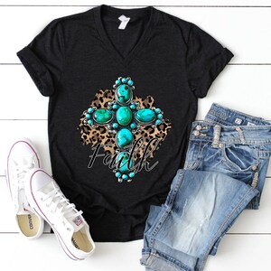 Puede incluir: Camiseta negra de cuello en V con un dise&ntilde;o de cruz turquesa y plata sobre un fondo de estampado de leopardo. La palabra "Faith" est&aacute; escrita debajo de la cruz.