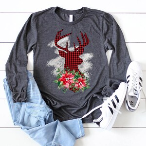 May include: A gray long-sleeved shirt with a red and black plaid deer silhouette design. The deer has a floral arrangement of red poinsettias and green leaves around its neck.