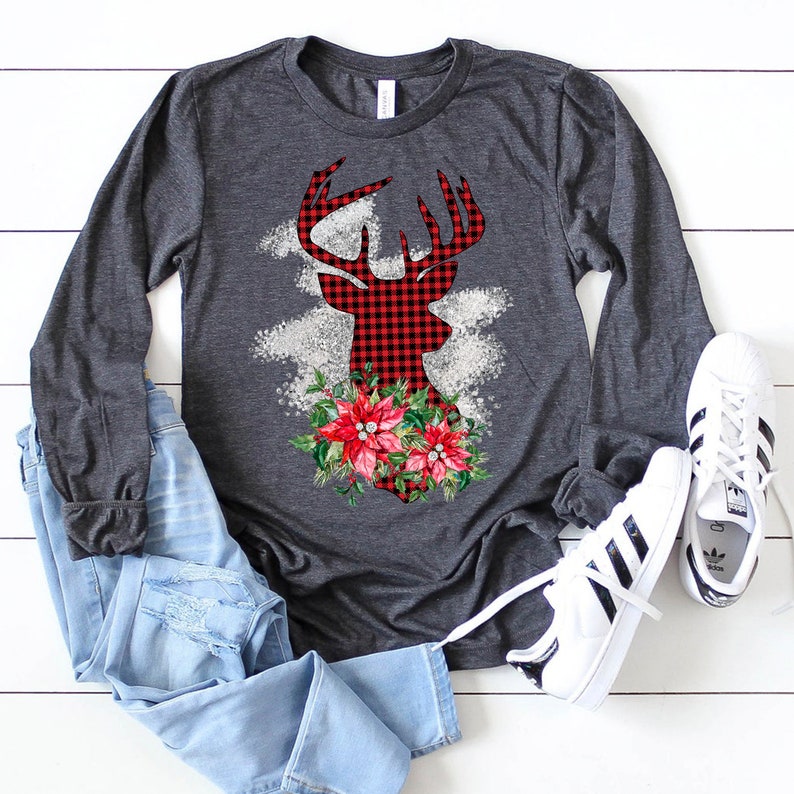 May include: A gray long-sleeved shirt with a red and black plaid deer silhouette design. The deer has a floral arrangement of red poinsettias and green leaves around its neck.