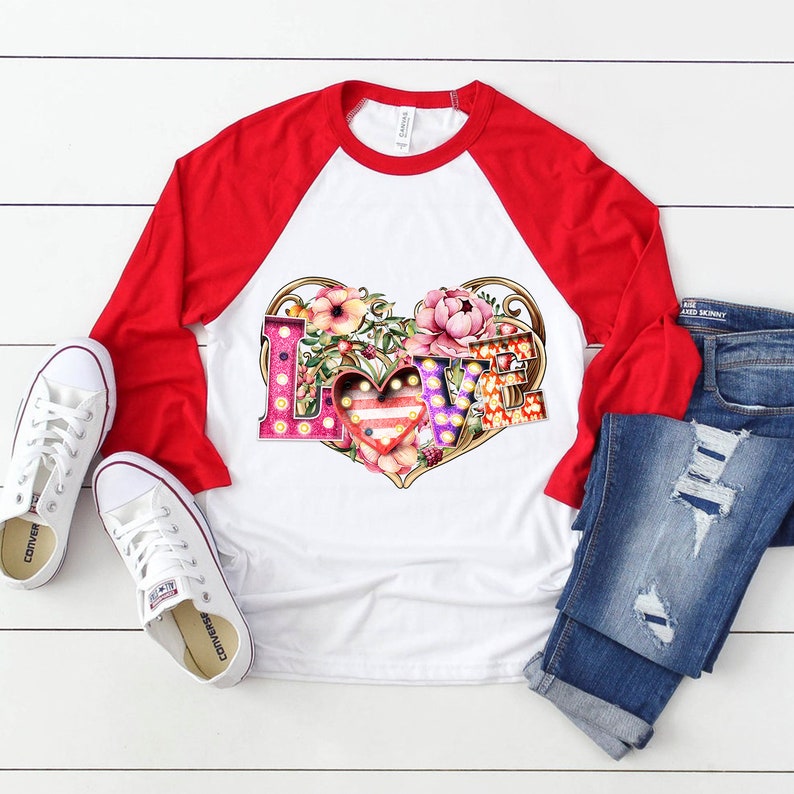 Puede incluir: Una camiseta de b&eacute;isbol blanca y roja con un gr&aacute;fico de la palabra "LOVE" en letras de colores con un coraz&oacute;n y flores. Las letras est&aacute;n decoradas con bombillas.