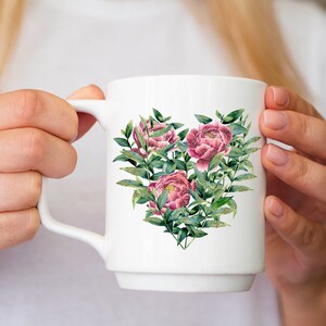 May include: White ceramic mug with a heart-shaped design featuring pink peonies and green leaves with gold glitter accents.