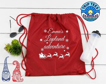 May include: A red drawstring backpack with white text that reads "Emma's Lapland adventure" with a reindeer and sleigh design. The bag has red drawstrings and is next to a water bottle, sunglasses, and gnome cutouts.