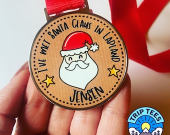 May include: A brown wooden medal with a gold rim and a cartoon image of Santa Claus. The medal says "I've met Santa Claus in Lapland Jensen".