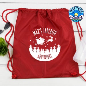 May include: Red drawstring bag featuring a white graphic of Santa Claus in a sleigh, reindeer, and trees, with the text "MAX'S LAPLAND ADVENTURE". The bag has red drawstrings and a Trip Tees logo.