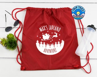 May include: Red drawstring backpack with a white graphic of Santa Claus in a sleigh, reindeer, and trees, with the text "MAX'S LAPLAND ADVENTURE". The bag has red drawstrings and a Trip Tees logo.