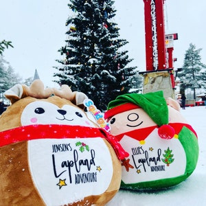 May include: Two plush toys, one a reindeer and one an elf, are sitting in the snow. The reindeer is brown with a red scarf and the elf is green with a red hat. Both toys have a white patch on their chest with the text "Lapland Adventure" and a name. The reindeer's name is Jensen's and the elf's name is Ellie's.