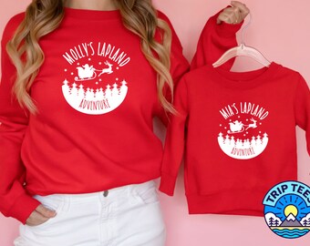 May include: Red sweatshirts with white graphic text. One sweatshirt reads "Molly's Lapland Adventure" and the other reads "Mia's Lapland Adventure". The graphic includes a Santa sleigh, reindeer, trees, and stars. The sweatshirts are a matching set.