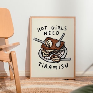 May include: A framed art print featuring a whimsical illustration of tiramisu with spoons. The text "HOT GIRLS NEED TIRAMISU" is written in black, playful lettering. The artwork is set against a cream-colored background, framed in a light wood.