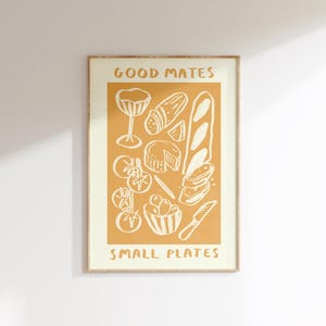 May include: A framed print with a mustard-colored background features white illustrations of food items. The words "GOOD MATES" and "SMALL PLATES" are at the top and bottom. The image includes a wine glass, bread, cheese, and tomatoes.