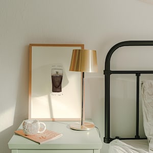 K&ouml;nnte beinhalten: Ein Nachttisch mit einem gerahmten Druck, einer Lampe und einer Tasse. Der Druck zeigt eine Zeichnung eines Guinness-Pints mit dem Text "Hap Giale Split the G". Eine goldfarbene Lampe mit zylindrischem Schirm steht auf dem Tisch.