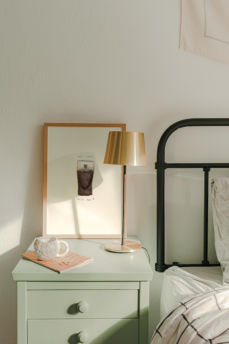 K&ouml;nnte beinhalten: Ein Nachttisch mit einem gerahmten Druck, einer Lampe und einer Tasse. Der Druck zeigt eine Zeichnung eines Guinness-Pints mit dem Text "Hap Giale Split the G". Eine goldfarbene Lampe mit zylindrischem Schirm steht auf dem Tisch.