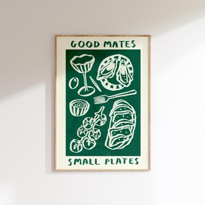 May include: A framed art print with a green background and white illustrations of food items. The text "GOOD MATES" and "SMALL PLATES" are at the top and bottom. The image includes a cocktail glass, olives, and a plate of food.