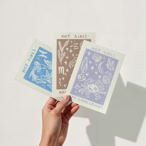 Puede incluir: Tres tarjetas con el texto "Hot Girls Are" seguido de un signo del zod&iacute;aco. La primera tarjeta es azul con Piscis, la segunda es marr&oacute;n con Escorpio y la tercera es morada con C&aacute;ncer.