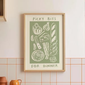 Könnte beinhalten: Ein gerahmter Druck mit grünem Hintergrund und beigen Illustrationen von Lebensmitteln, darunter Oliven, Käse, Brot und eine Feige. Der Text "Picky Bits For Dinner" ist in Beige dargestellt. Der Druck befindet sich in einem hellbraunen Holzrahmen.