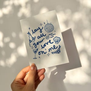 May include: A white card with blue ink illustrations of a disco ball, a cocktail glass, a seashell, a beach umbrella, and a party hat. The text on the card reads "Lay all your love on me".
