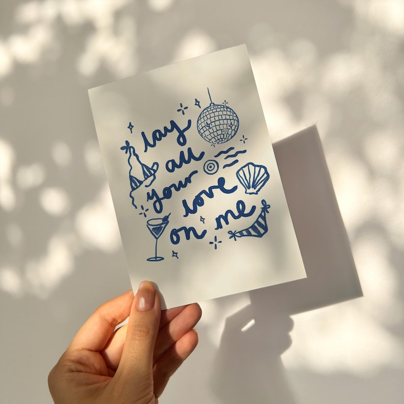 May include: A white card with blue ink illustrations of a disco ball, a cocktail glass, a seashell, a beach umbrella, and a party hat. The text on the card reads "Lay all your love on me".