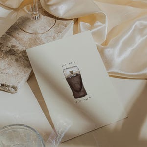 K&ouml;nnte beinhalten: Cremefarbene Gru&szlig;karte mit einer Aquarellillustration eines Guinness-Pints. Auf der Karte steht "Hot Girls Split the G". Ein Glas und ein Marmorblock sind ebenfalls im Bild zu sehen.